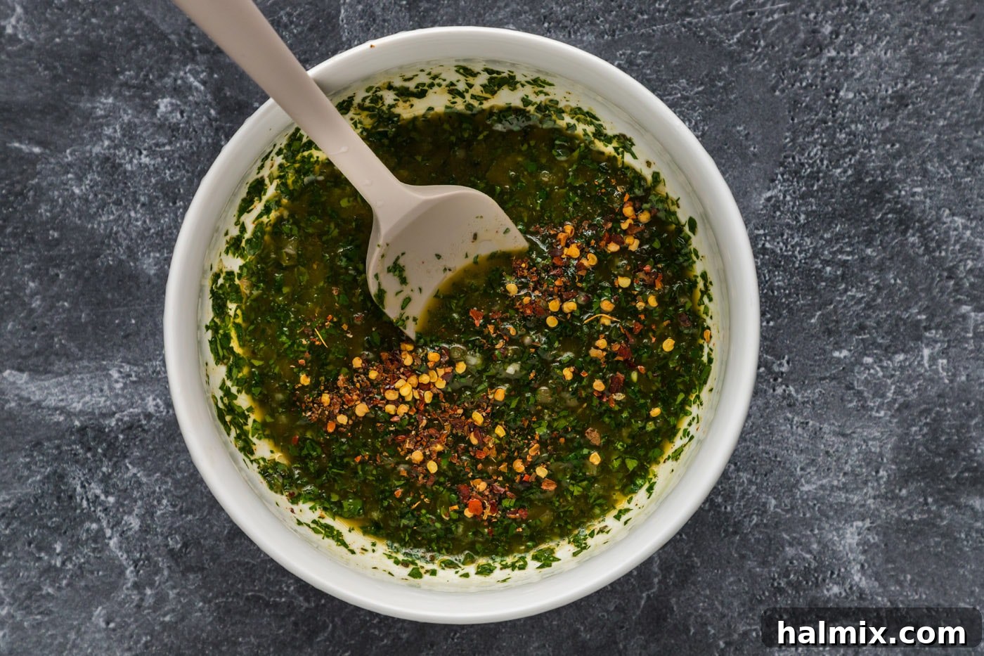 Adding Red Pepper Flakes to Chimichurri Fresh chimichurri sauce in a mixing bowl, being mixed with red pepper flakes.