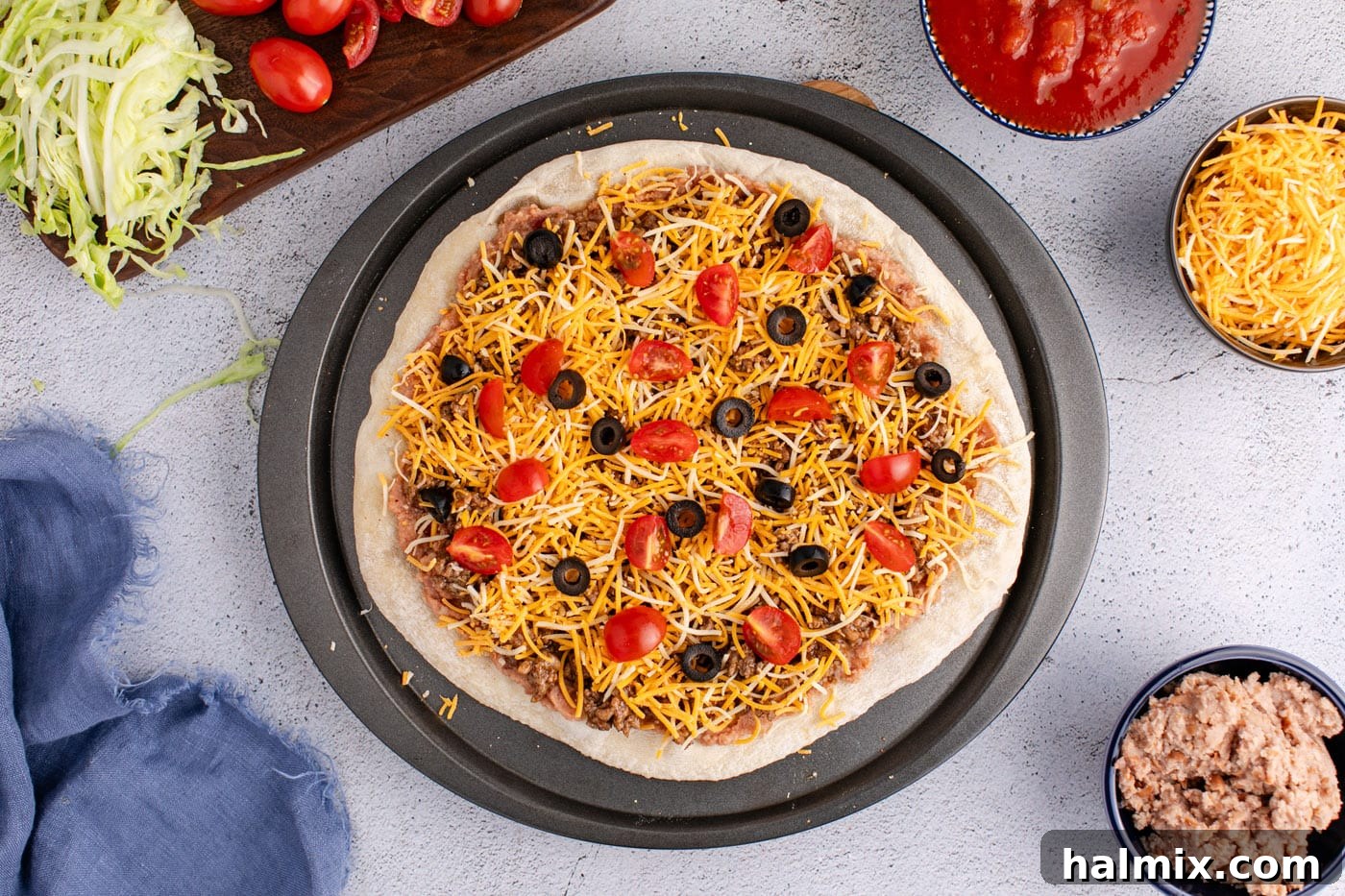 Shredded cheese, taco meat, tomatoes, and olives added on top of refried beans and pizza crust before baking