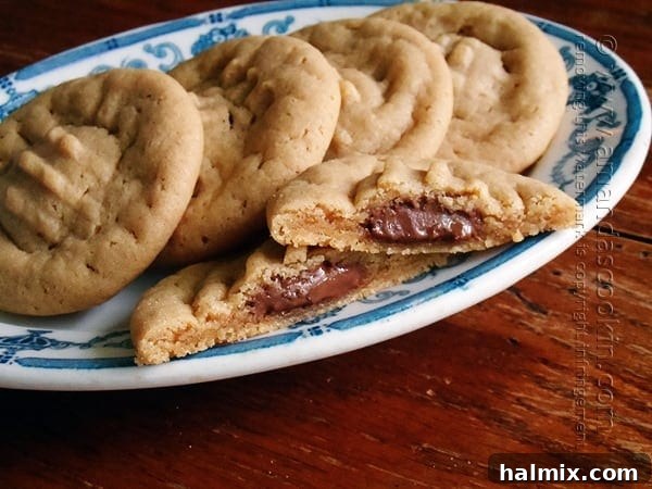 Melting Chocolate Core Peanut Butter Cookies 2 A close-up of a Hershey drop stuffed peanut butter cookie, perfectly split in half on a plate, revealing its warm, melted chocolate center. Whole cookies are blurred in the background, hinting at more deliciousness.