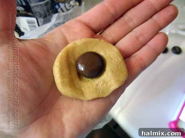 Melting Chocolate Core Peanut Butter Cookies 5 A close-up view of a hand gently flattening a ball of peanut butter cookie dough in the palm, with a single Hershey's Drop chocolate placed precisely in the center, ready to be wrapped.