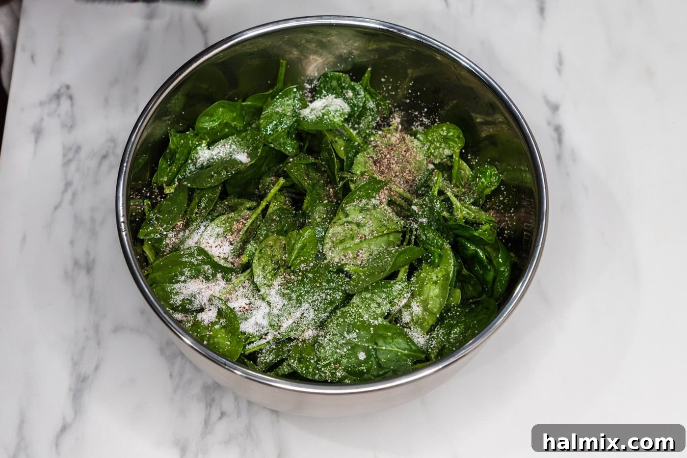 Spinach leaves in a bowl with salt, pepper, and olive oil, being tossed to season.