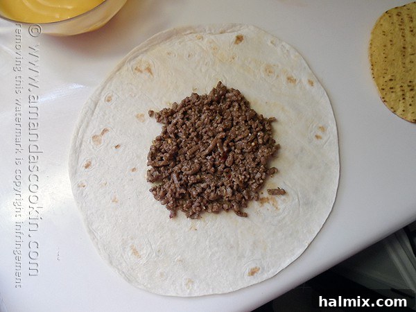 An overhead photo of a large flour tortilla with cooked and seasoned ground beef spread in the center, ready for the next layer of the Crunchwrap Supreme.