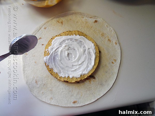 Sour cream being spread evenly over the top of a tostada shell, which sits atop the seasoned ground beef and nacho cheese in a flour tortilla for a homemade Crunchwrap.
