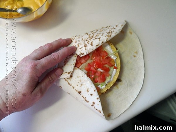 The edges of a large flour tortilla being carefully folded inward and creased around the central layers of a Crunchwrap Supreme, preparing it for grilling.