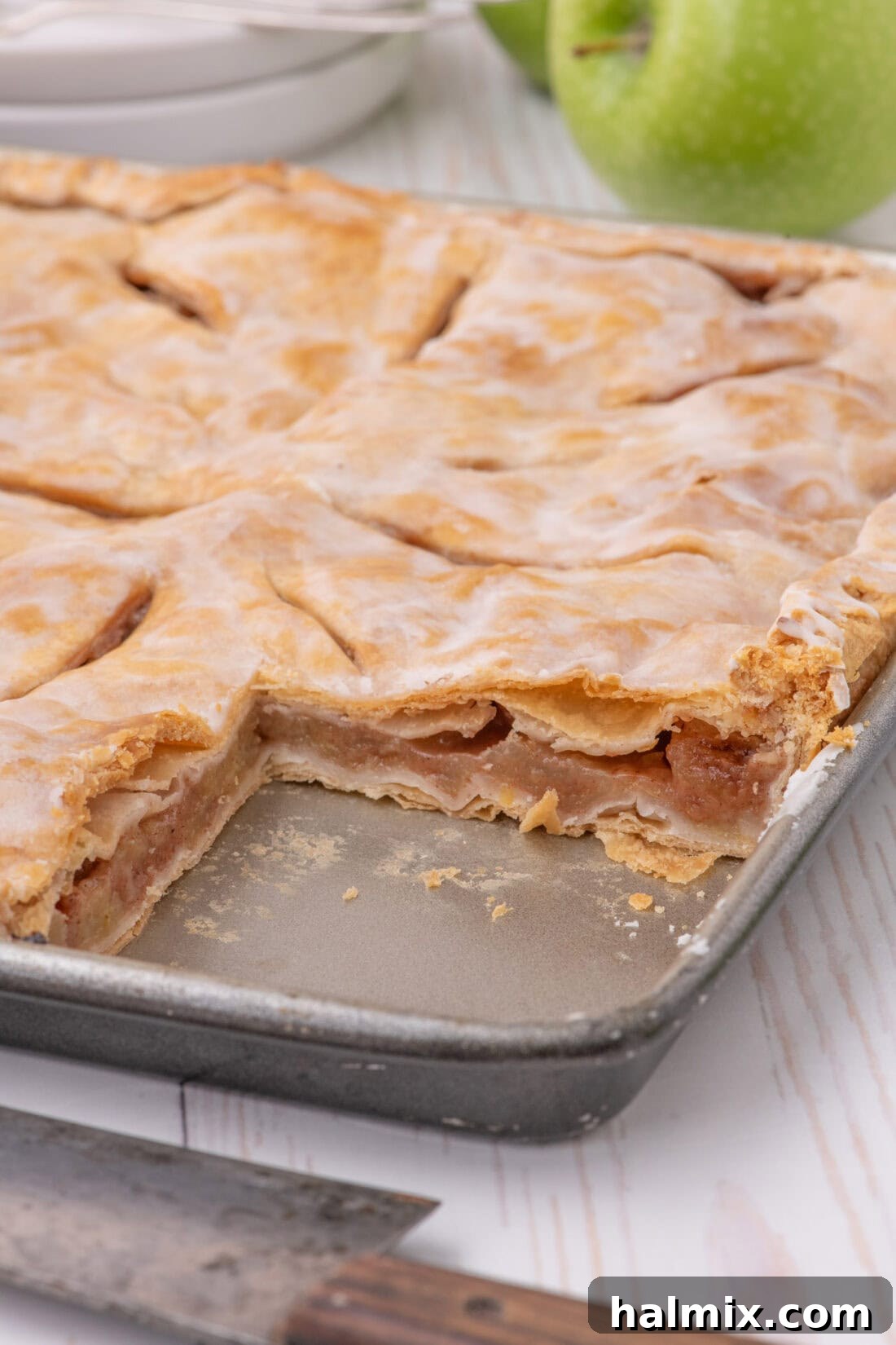 A large rectangular pan of baked Apple Slab Pie with one corner slice neatly removed, showcasing the abundant apple filling and a beautifully glazed crust, inviting guests to enjoy.
