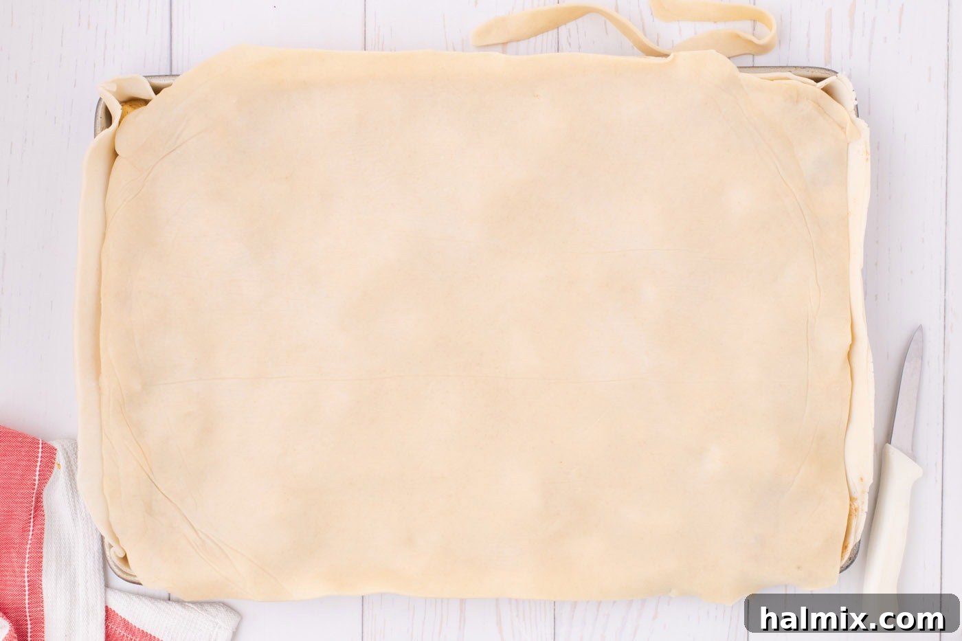 The top pie crust layer covering the apple filling in the jelly roll pan, with excess dough still to be trimmed and sealed.