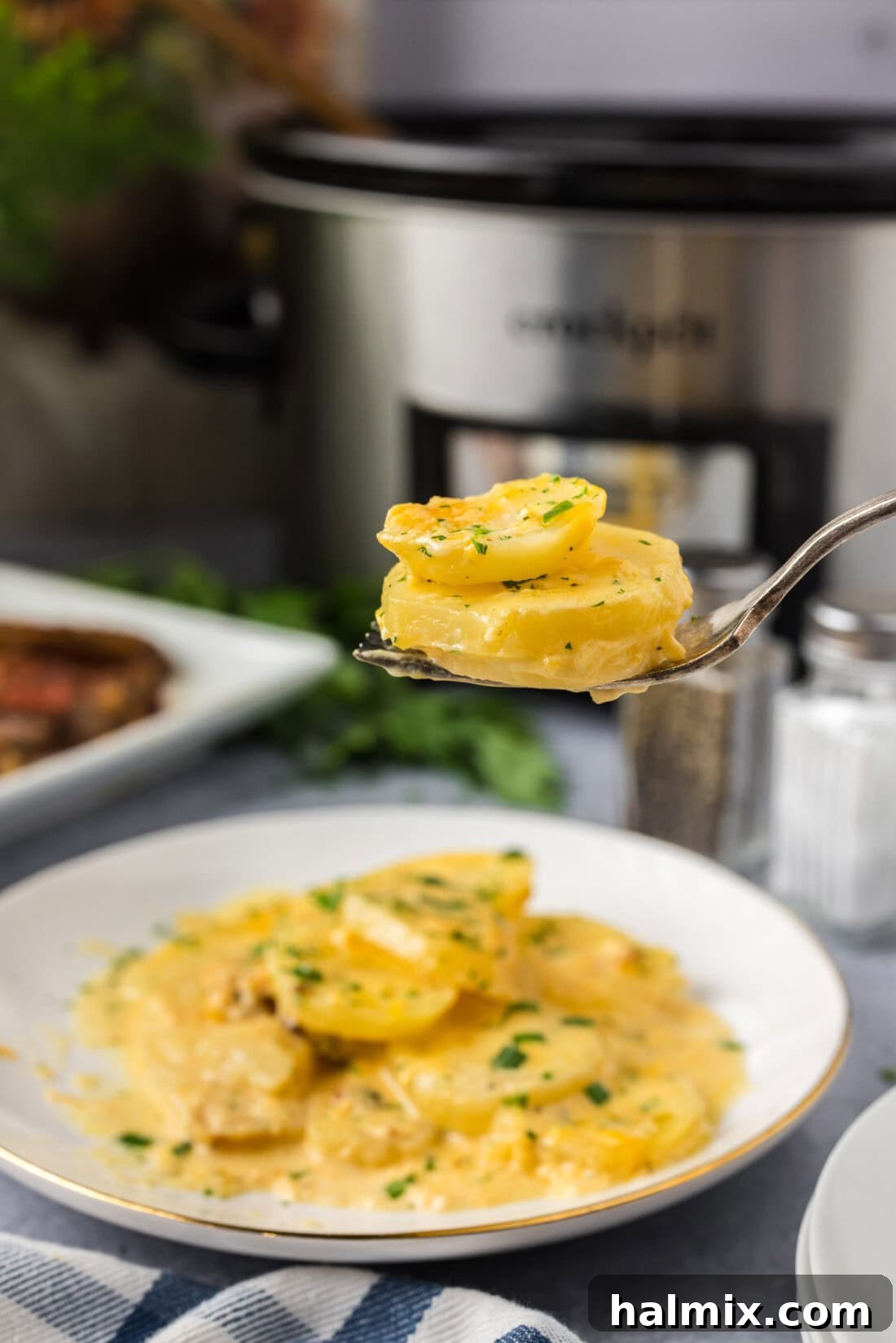 Rich and Creamy Slow Cooker Scalloped Potatoes 2 Two Crockpot Scalloped Potatoes on a fork held above a plate of Crockpot Scalloped Potatoes