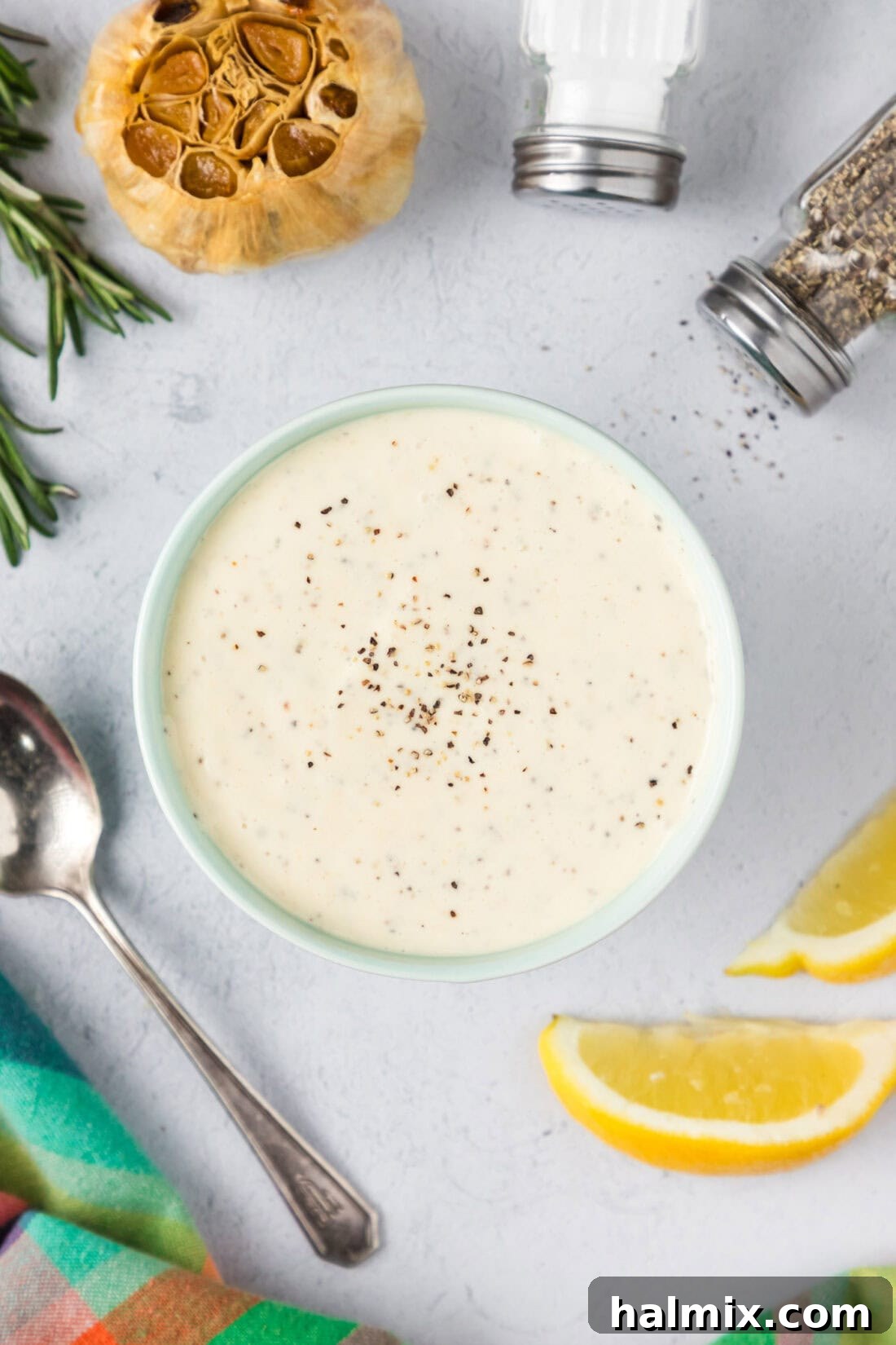 A beautifully presented bowl of creamy Roasted Garlic Aioli, garnished with fresh herbs, alongside lemons, whole garlic cloves, and seasonings, creating an inviting culinary scene.