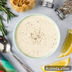 Close up photo of a bowl of creamy homemade Garlic Aioli with fresh lemons, whole garlic cloves, and seasonings scattered around it on a rustic surface.