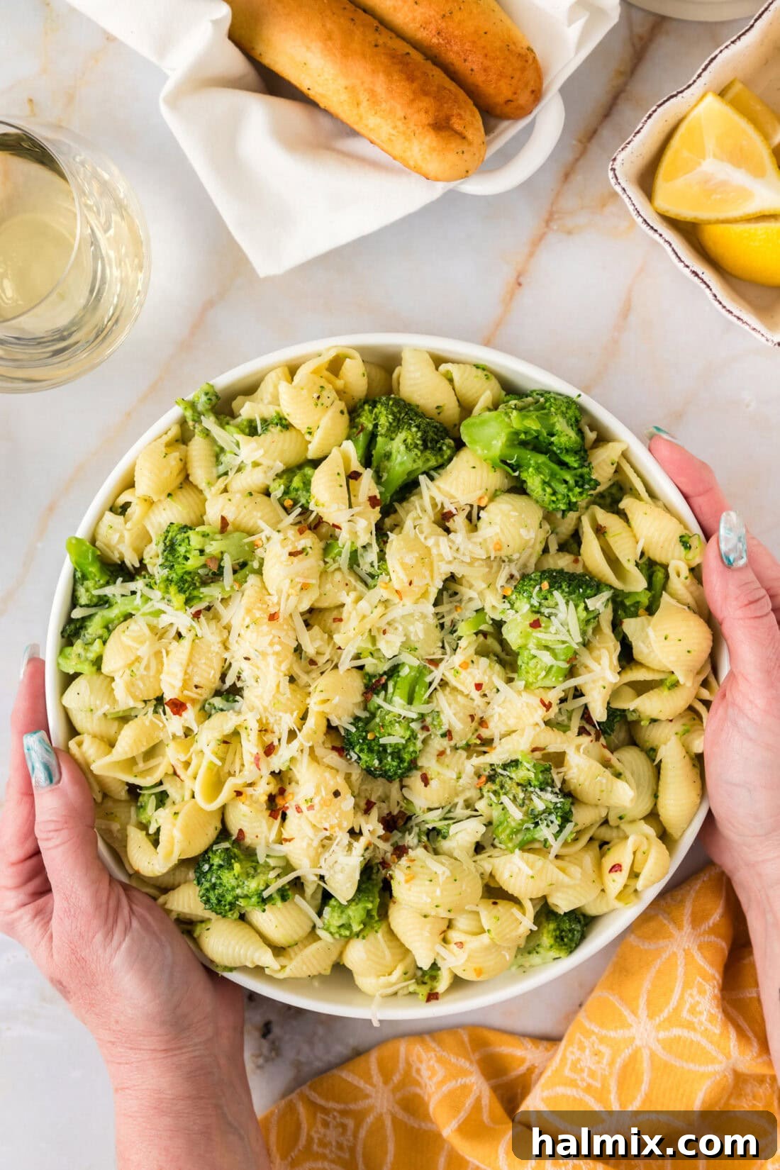 Hands holding a bowl of Broccoli Pasta, garnished with red pepper flakes and a lemon wedge.