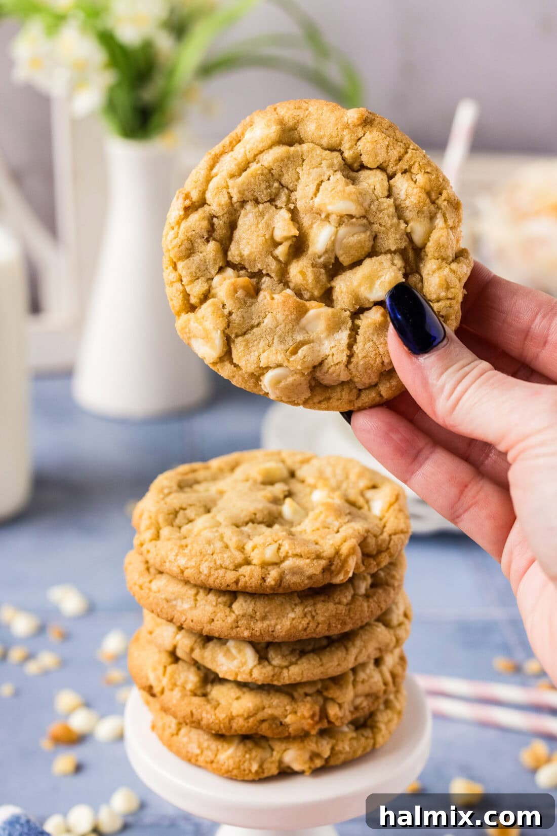 A hand holding up a freshly baked White Chocolate Macadamia Nut Cookie, with a stack of more delicious cookies blurred in the background, highlighting its golden perfection.