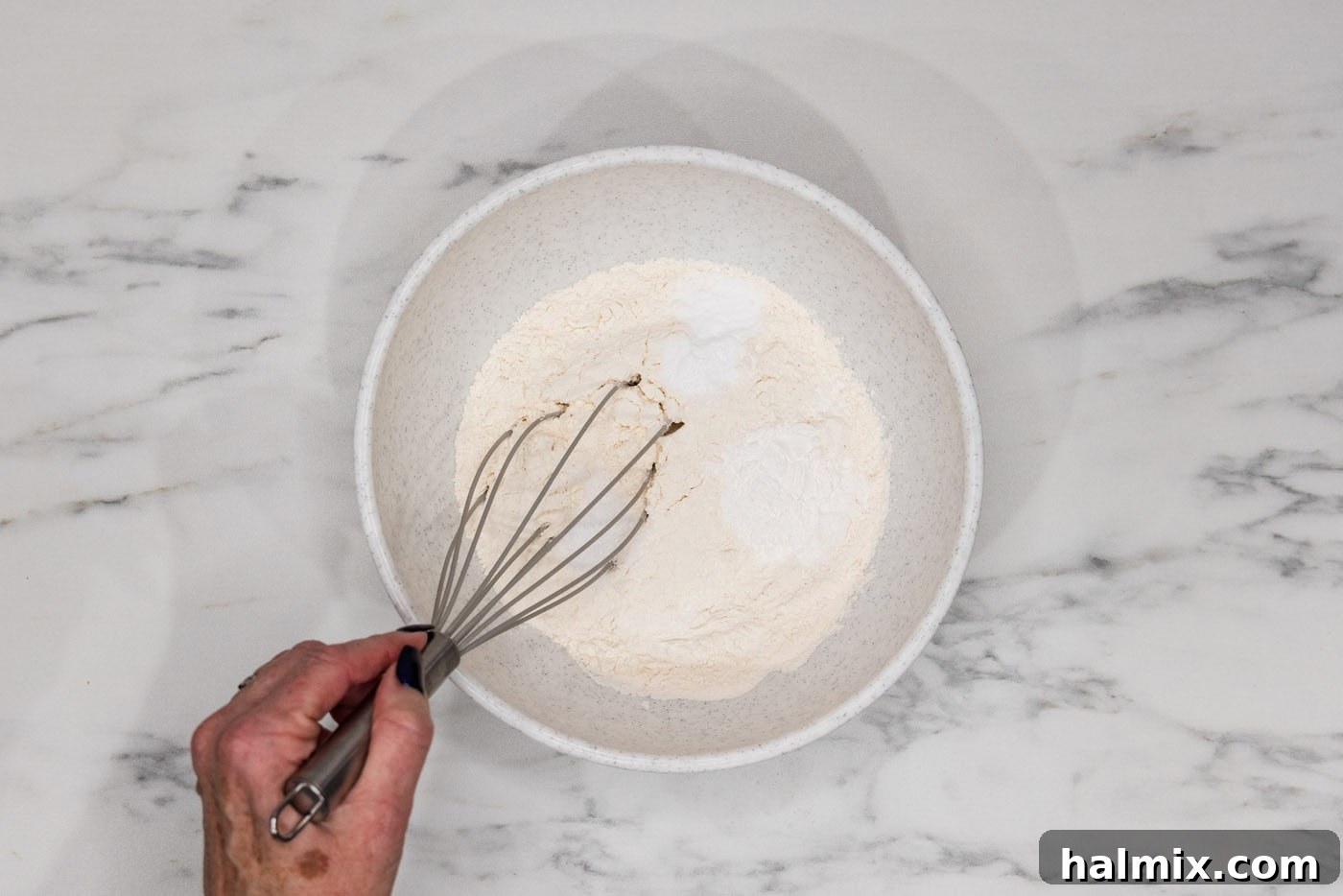 A hand whisking together flour, baking powder, baking soda, and salt in a bowl, preparing the dry ingredients for the cookie dough.