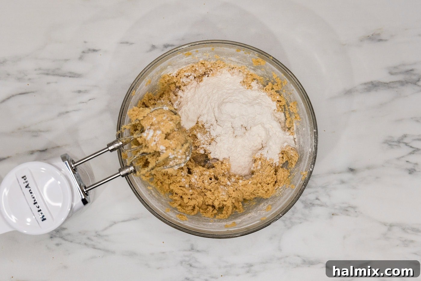 A hand mixer blending dry ingredients into wet cookie dough in a bowl, showing the gradual mixing process.