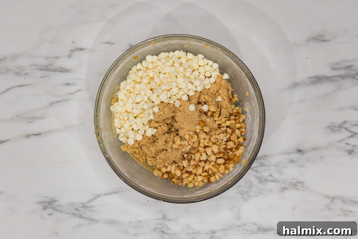 White chocolate chips and macadamia nuts have just been added to the cookie dough in a bowl, ready to be folded in.