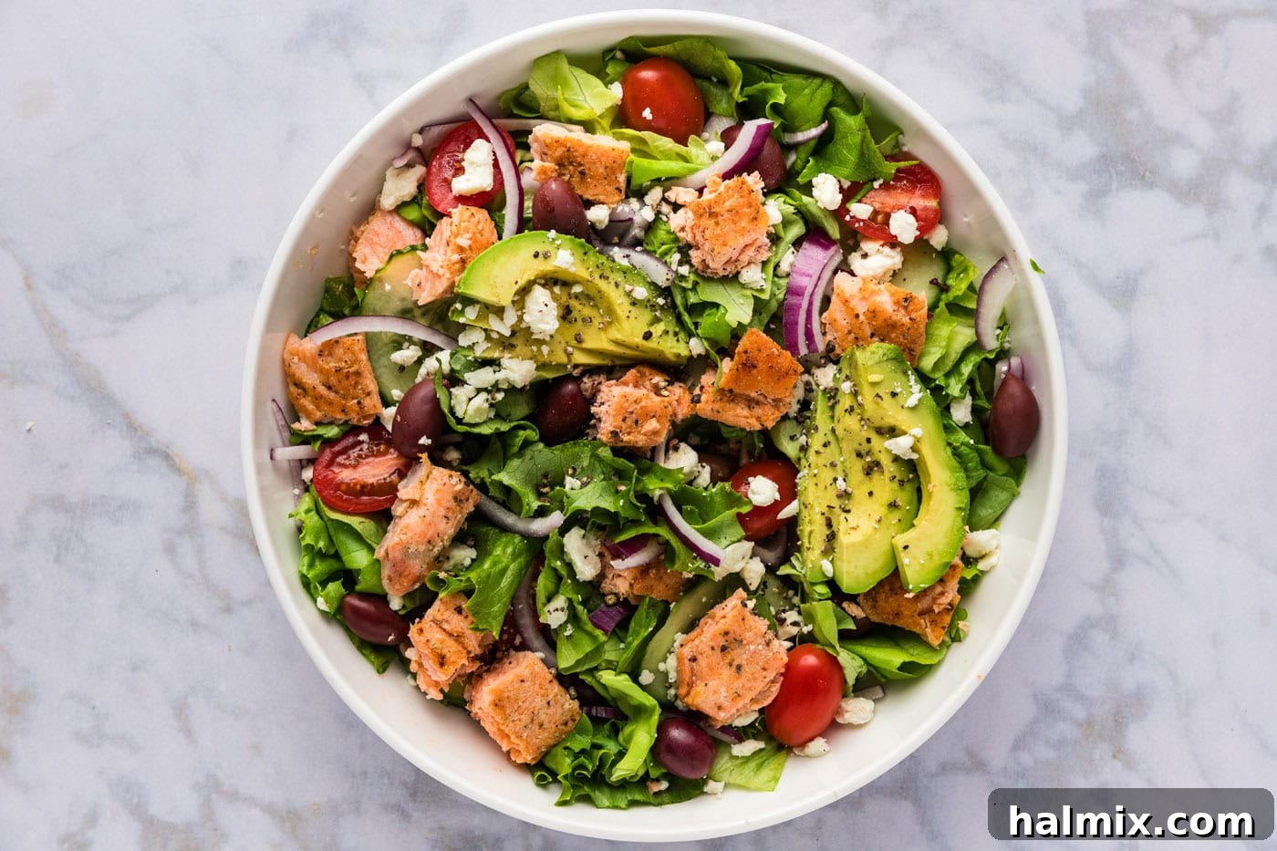 Close-up of a salad bowl with avocado slices, crumbled feta cheese, and cooked salmon cubes added on top of the mixed greens