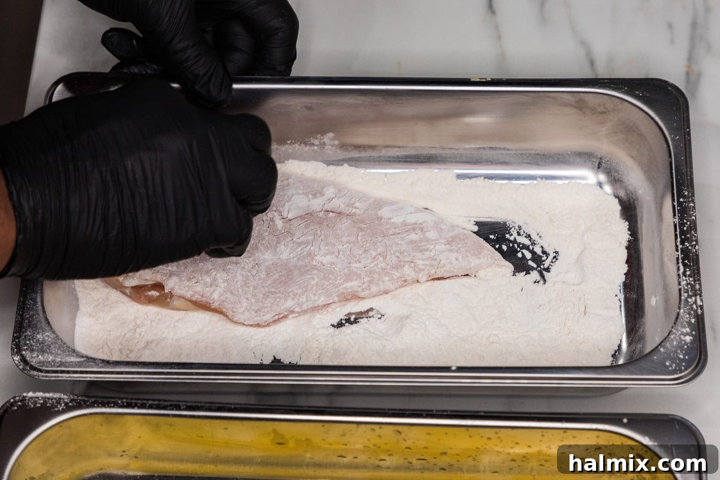 A hand dipping a chicken breast into a tray of flour mixture.