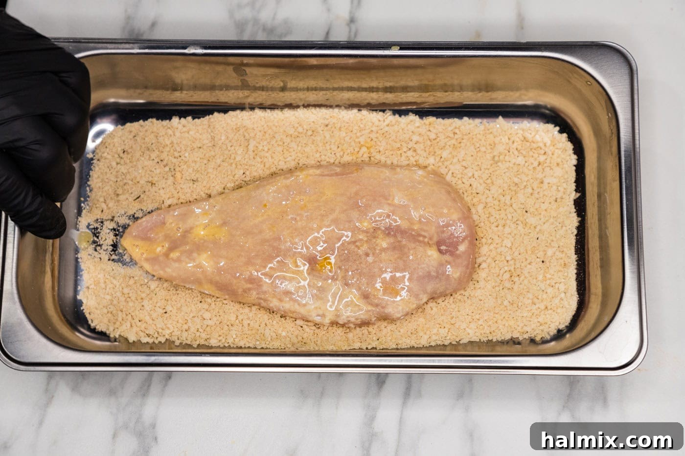 A breaded chicken breast nestled in a pan filled with panko breadcrumbs, showing full coverage.