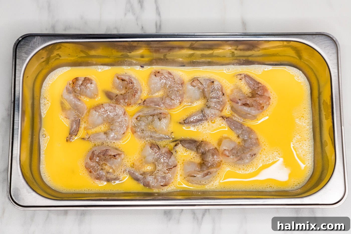 Shrimp being coated in whisked egg in a breading tray.