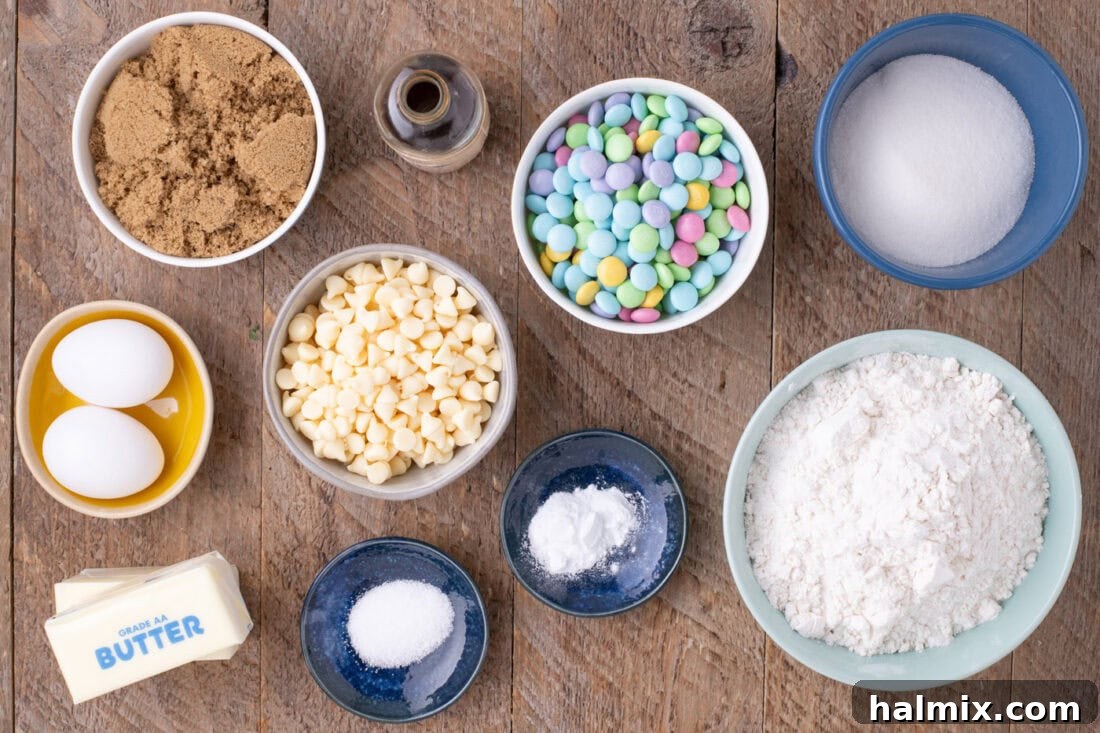 A selection of ingredients for Easter M&M Cookies laid out on a surface, including flour, sugars, butter, eggs, vanilla, white chocolate chips, and pastel M&Ms.