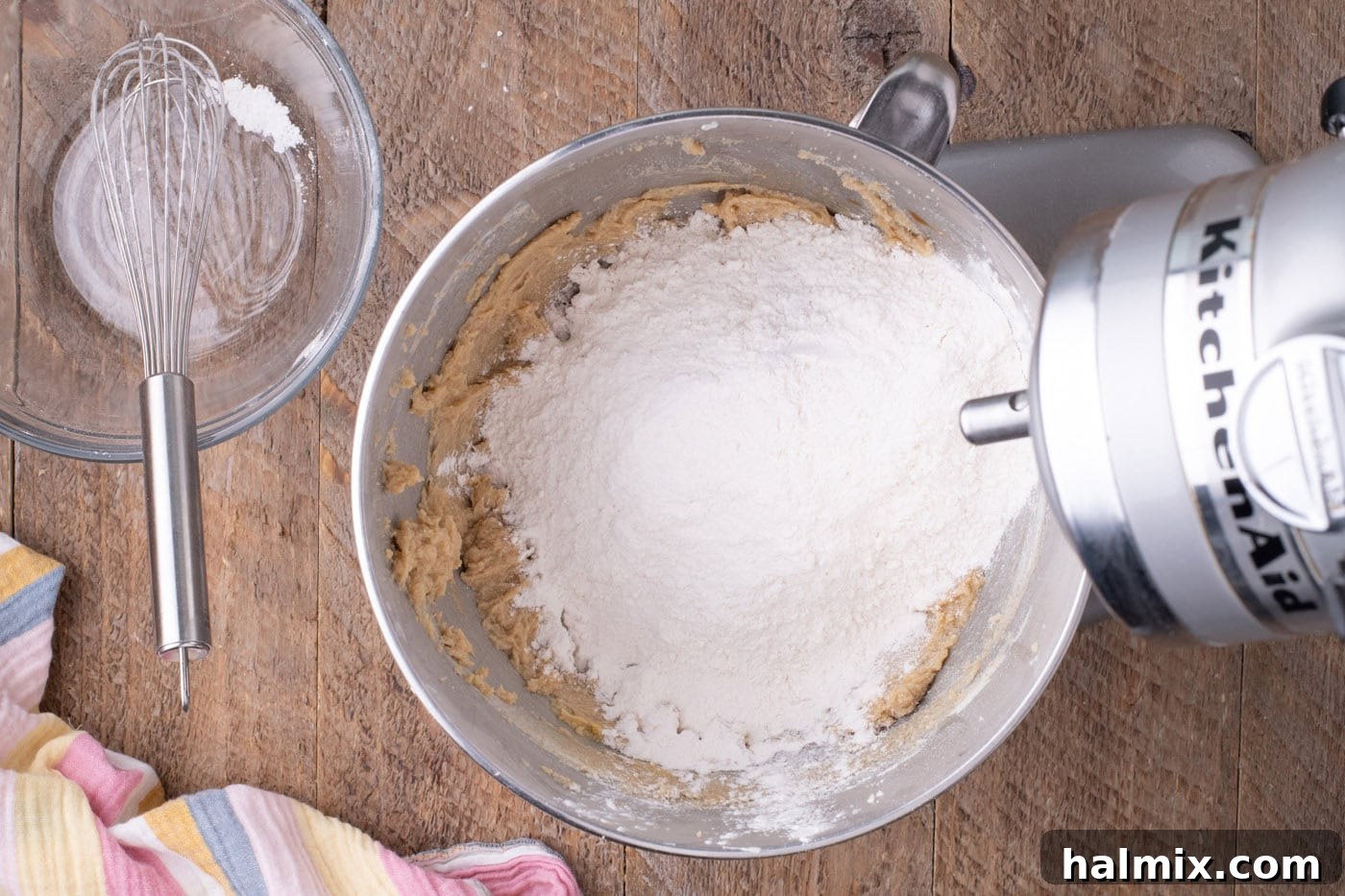 Dry ingredients gradually added to the wet ingredients in a stand mixer bowl, forming cookie dough.