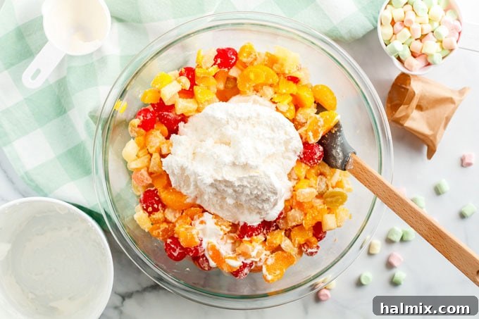 A step-by-step image showing Cool Whip being added over a bowl of fruit cocktail, ready to be mixed into Ambrosia Salad.
