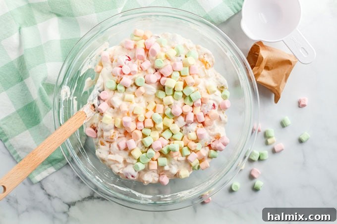 A vibrant display of colorful miniature marshmallows delicately folded over a fruit cocktail mixture, an essential final step for Ambrosia Salad.