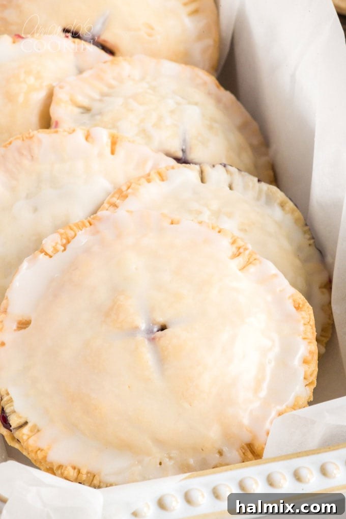 Making blueberry hand pies is fun and easy, and using a refrigerator pie crust dough cuts down on the work. Mini blueberry pies are delicious! A captivating close-up of several glazed blueberry hand pies neatly arranged in a rustic dish, showcasing their glistening topping.