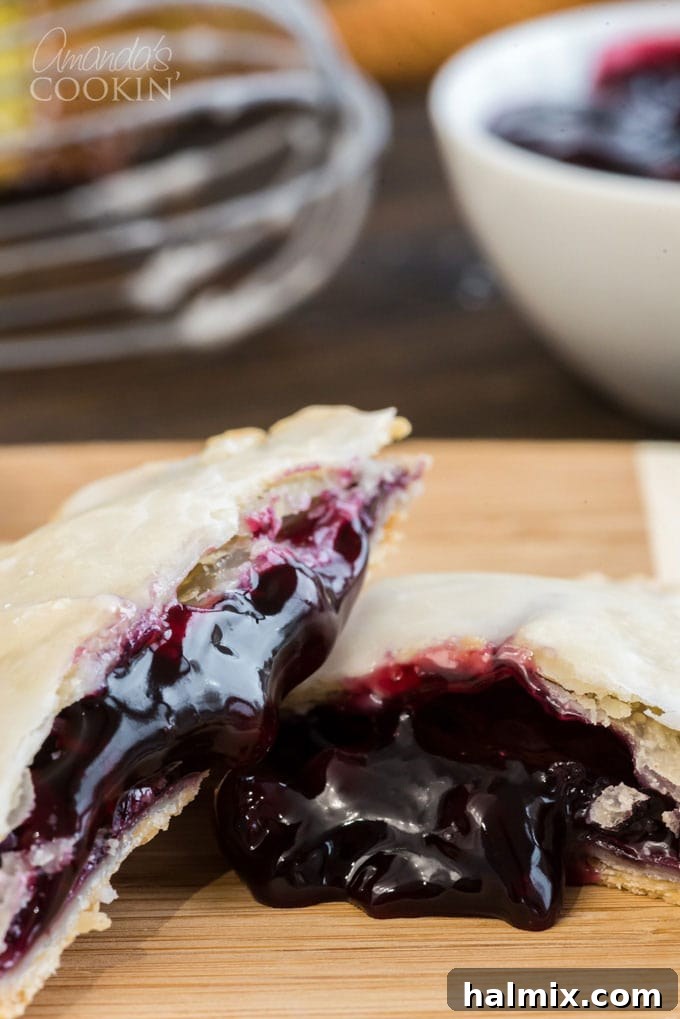 Making blueberry hand pies is fun and easy, and using a refrigerator pie crust dough cuts down on the work. Mini blueberry pies are delicious! A freshly baked hand pie, elegantly broken in half, showcasing the vibrant, juicy blueberry filling spilling out.