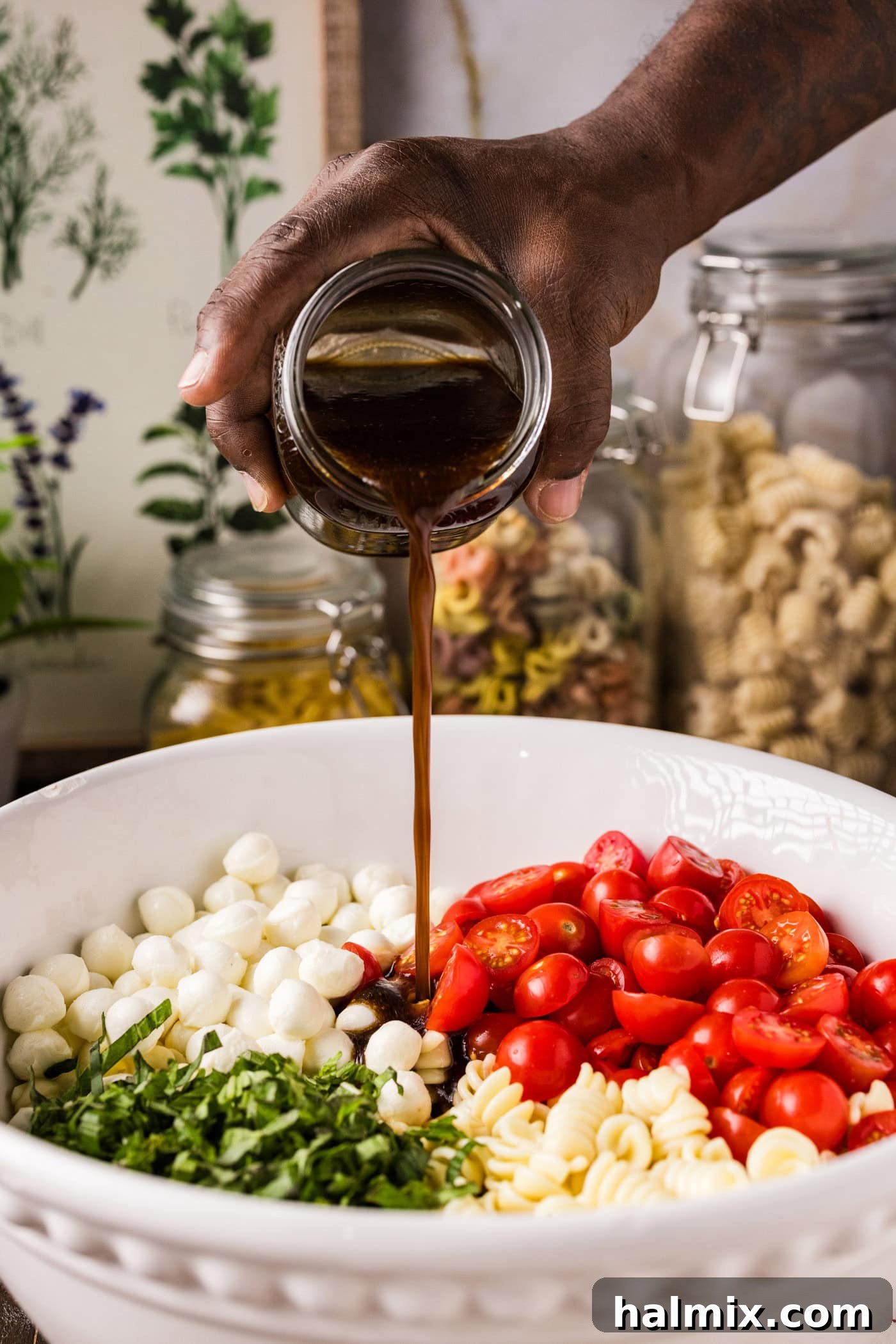 Pouring homemade balsamic vinaigrette generously over the Caprese Pasta Salad ingredients in a mixing bowl
