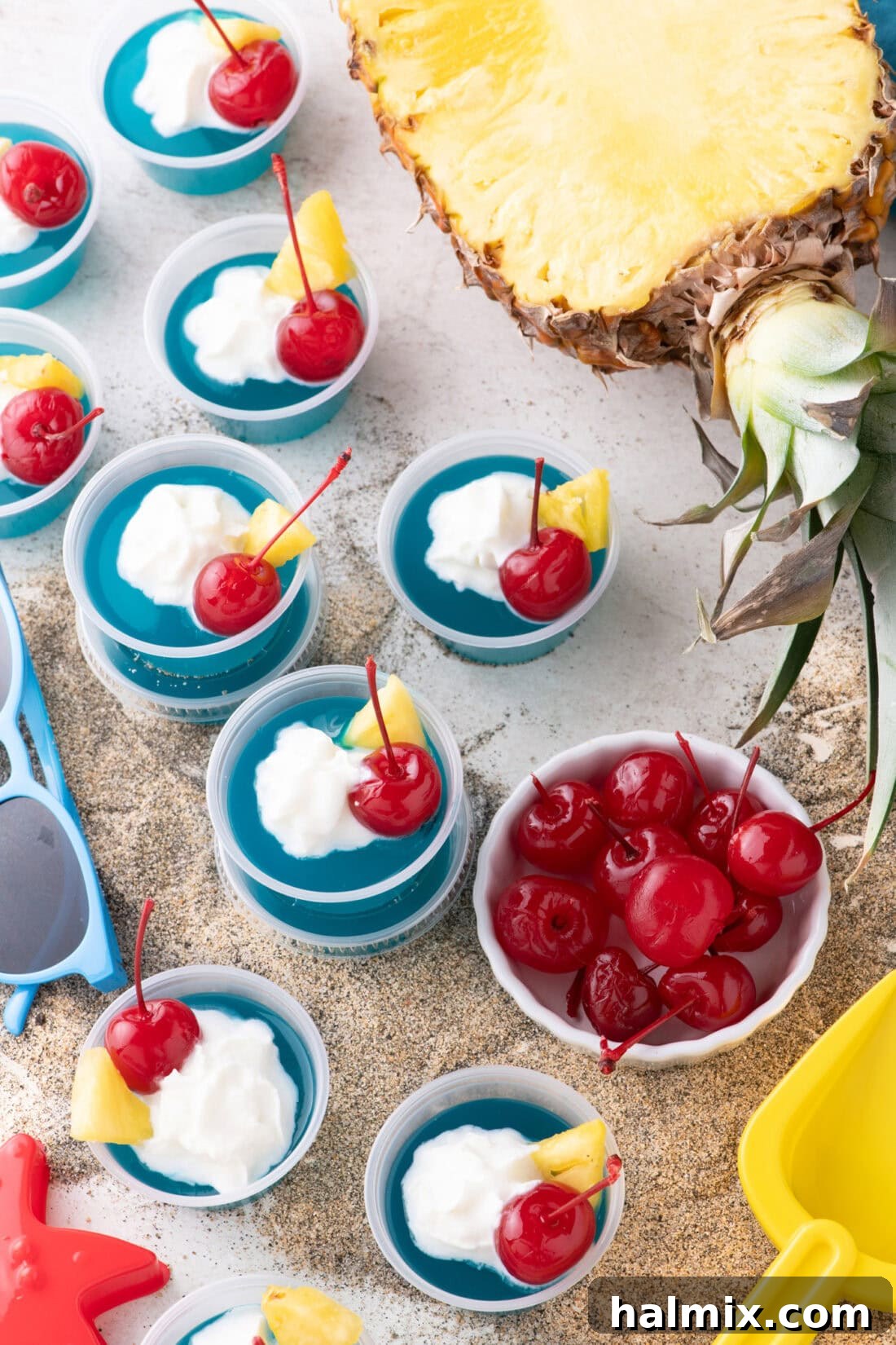 Blue Hawaiian Jello Shots resting on sand with a pineapple and cherries in the background