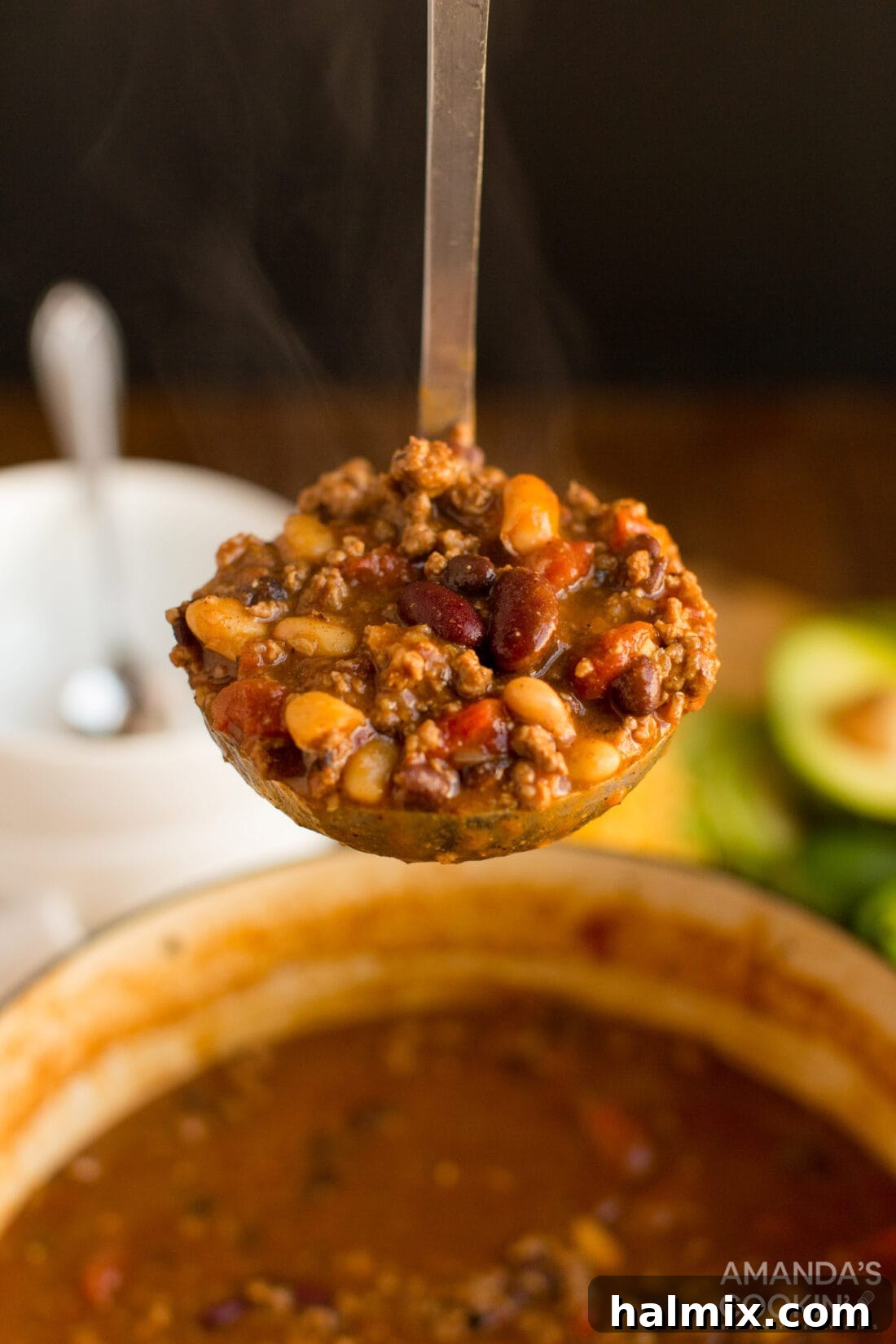A ladle full of rich, three-bean chili, garnished with fresh cilantro and shredded cheese, ready to be served into a bowl.