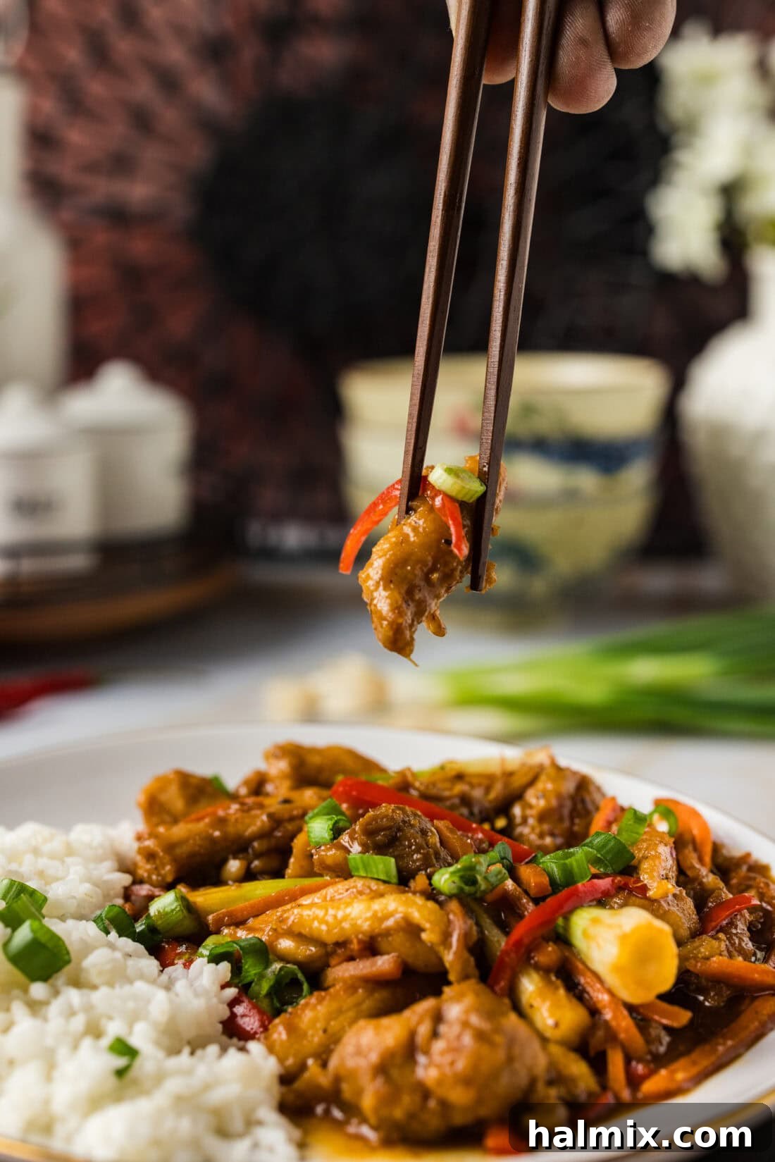 A chopstick holds a succulent piece of Szechuan Chicken over a generous plate of the finished dish, inviting diners to savor its rich flavors and textures.