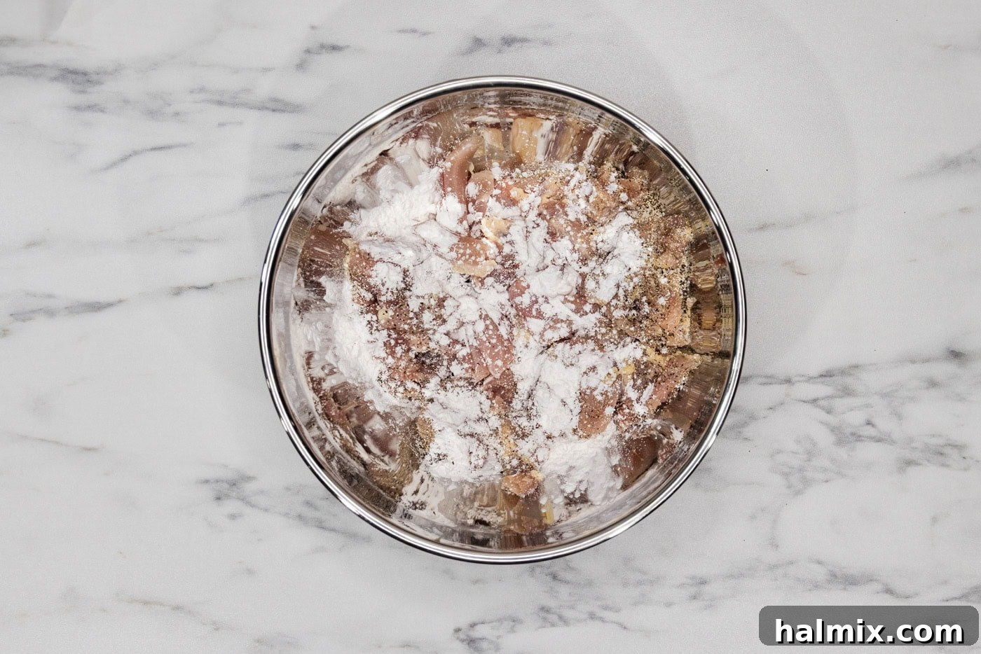 Sliced chicken in a bowl with cornstarch sprinkled on top, ready for coating.