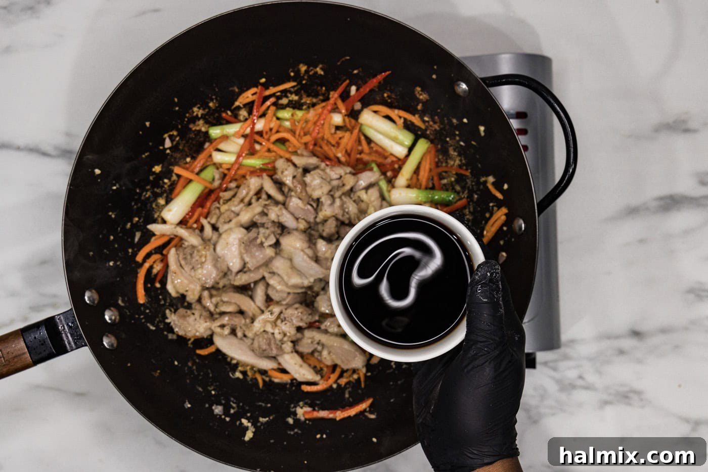 A gloved hand pouring soy sauce from a bowl into a skillet filled with chicken and stir-fried vegetables, ready to be sauced.