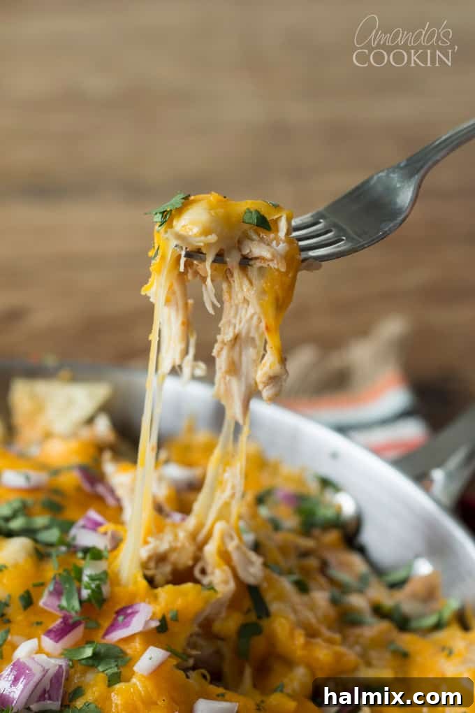 A close-up photo of a fork pulling a piece of cheesy rotisserie chicken nachos from a pan, highlighting the melted cheese pull.