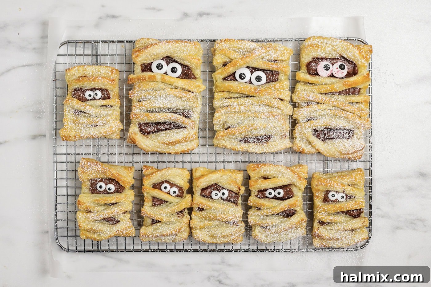 Powdered sugar being dusted over mummy hand pies on a wire cooling rack, creating a ghostly effect