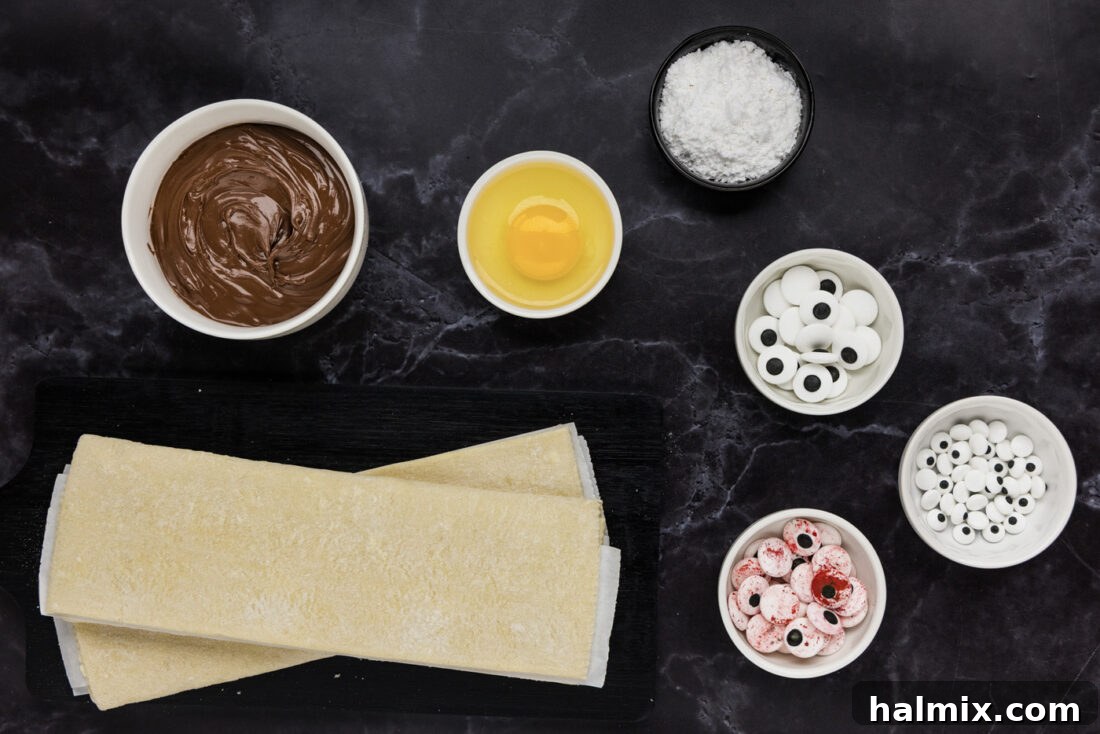 Ingredients for Mummy Hand Pies laid out on a white surface, including puff pastry, Nutella, jam, an egg, and candy eyeballs