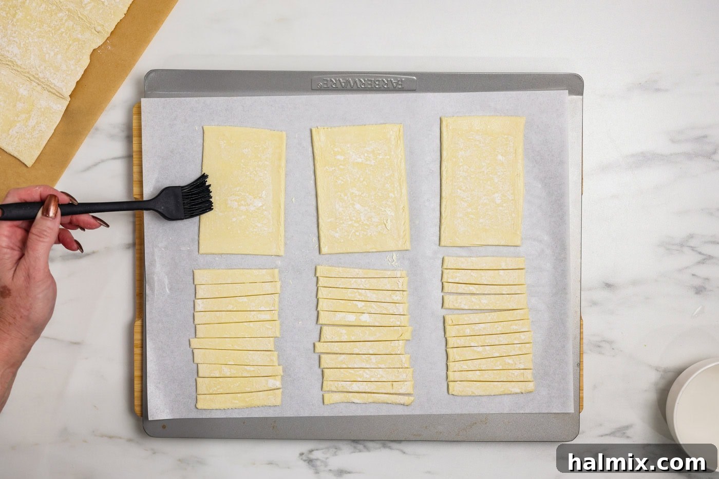 A hand using a pastry brush to lightly moisten the edges of a rectangular puff pastry piece with water
