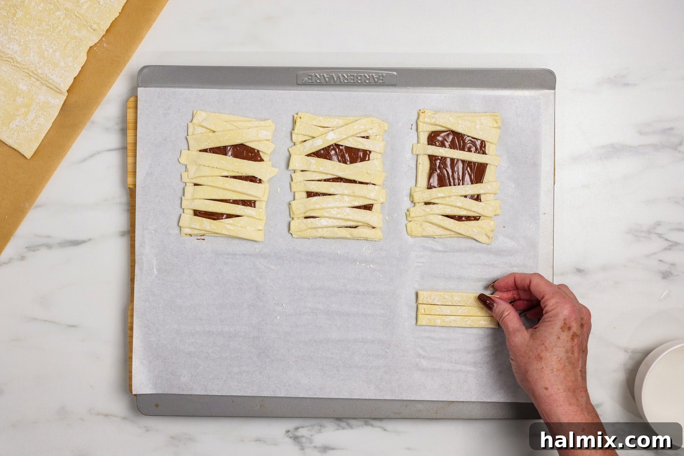 A hand carefully placing thin strips of puff pastry over a Nutella-coated rectangle to form mummy bandages
