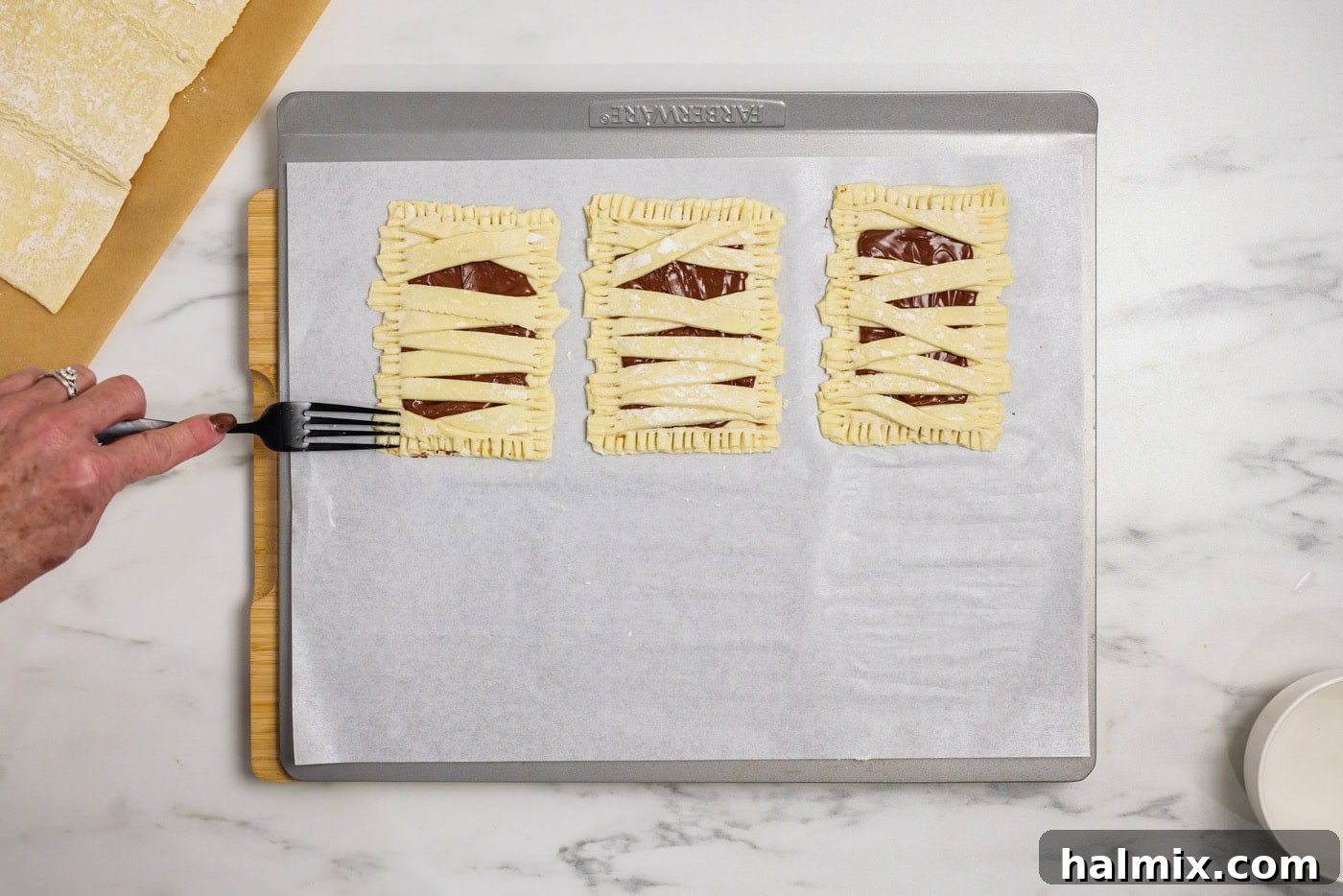 A fork pressing into the edges of assembled mummy hand pies to seal them securely before baking