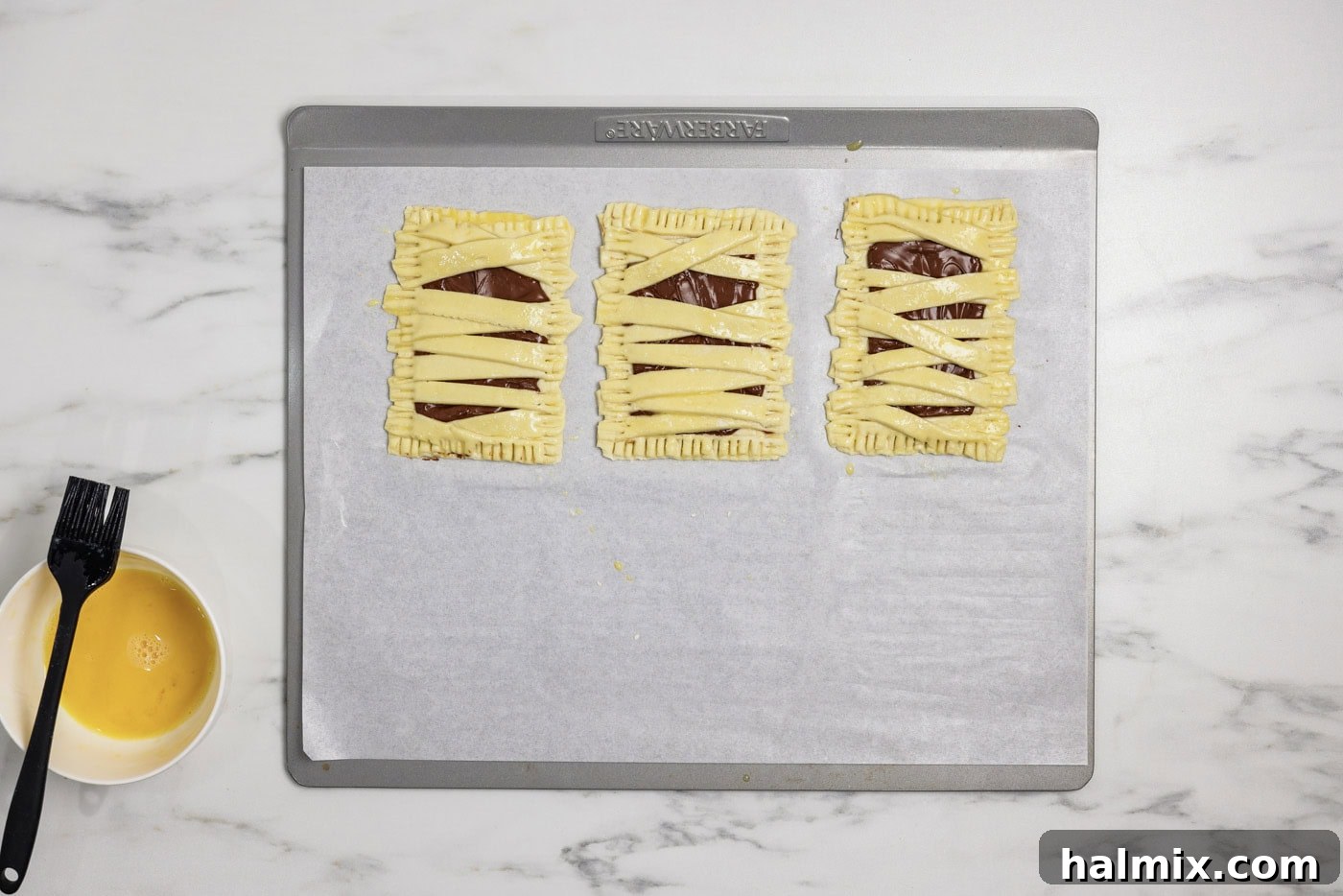 Mummy hand pies brushed with egg wash resting on a parchment-lined baking sheet, ready for the oven