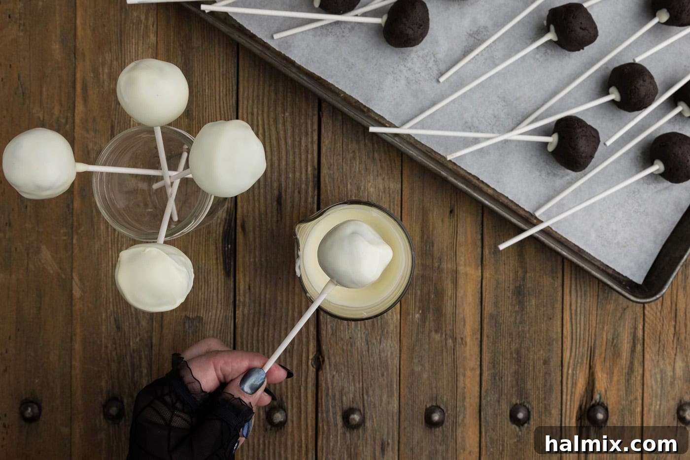 A hand carefully dipping an Oreo truffle, held by a lollipop stick, into a bowl of smooth, melted white chocolate coating.