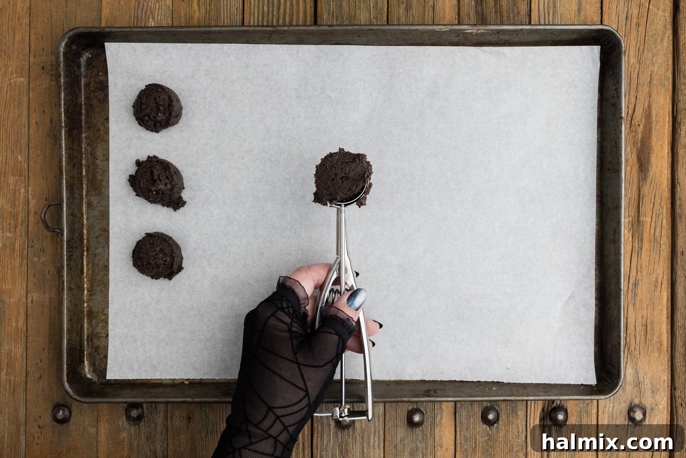 A 1-tablespoon cookie scoop holding a portion of the Oreo truffle mixture, poised above a parchment-lined baking sheet, demonstrating the first step in forming the balls.