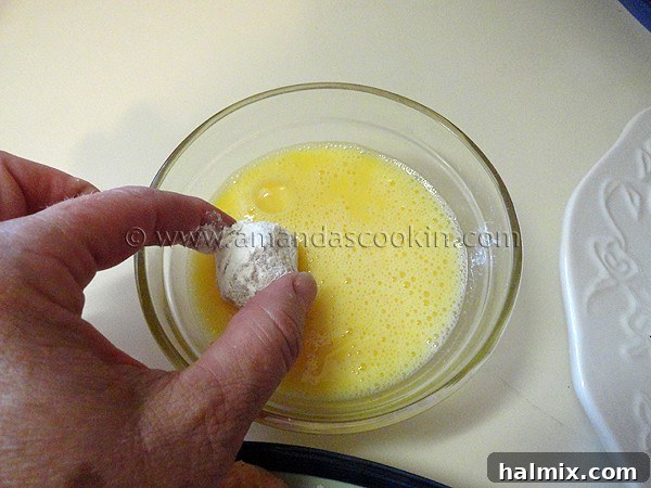 A flour-coated chicken ball being dipped into the bowl of whisked eggs, ensuring a complete and even liquid coating.