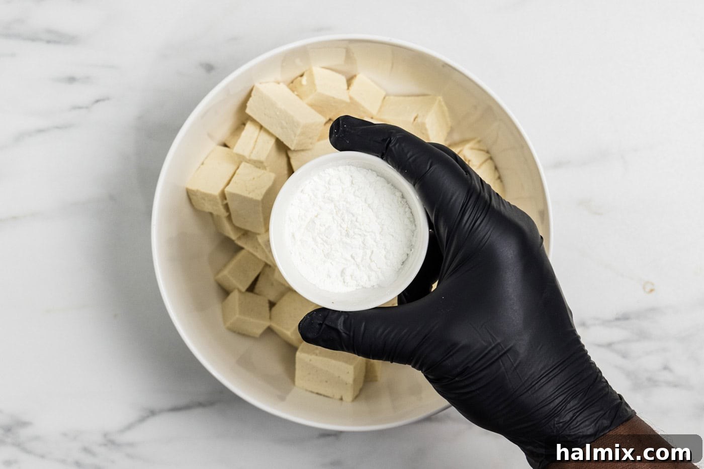 Golden Tofu Stir-Fry 5 Hand holding a bowl of cornstarch over cubed tofu in a bowl, demonstrating the coating process.