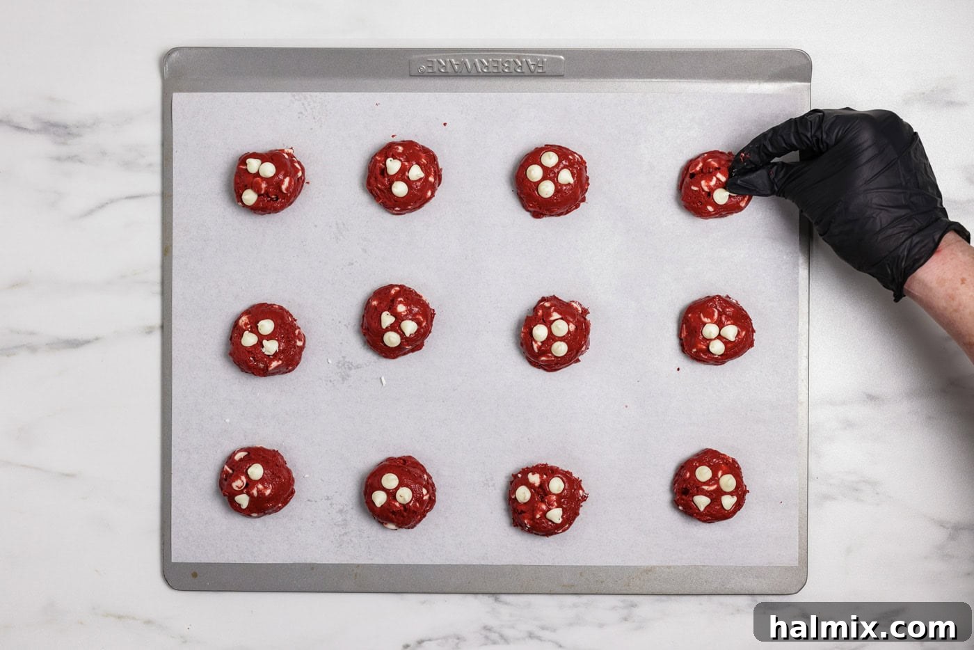hand dotting red velvet cookie dough balls with white chocolate chips on a baking sheet