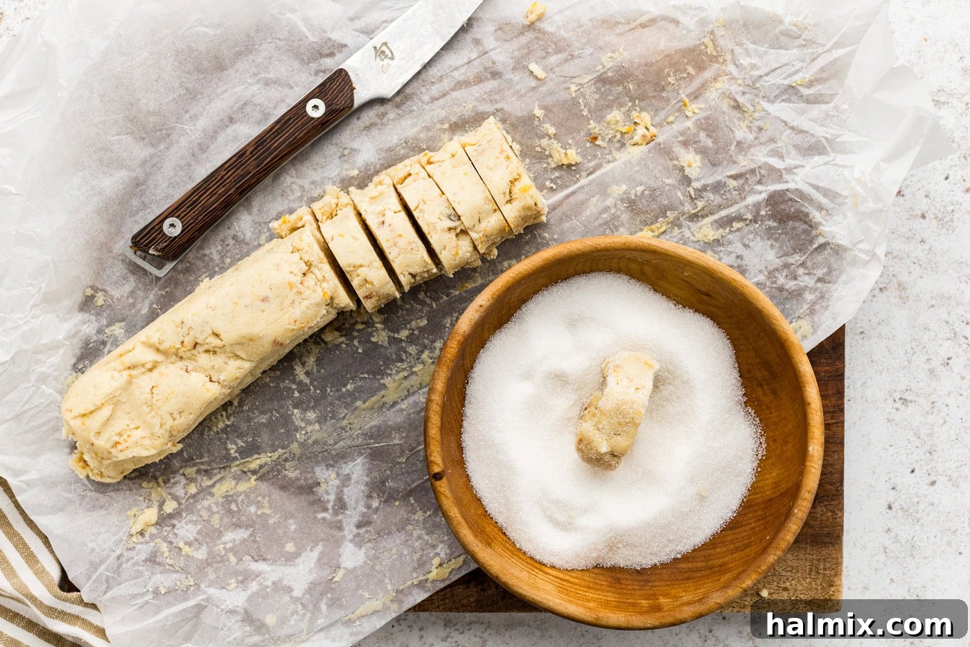 Sliced icebox cookie dough being rolled in a bowl of granulated sugar for a sweet coating.