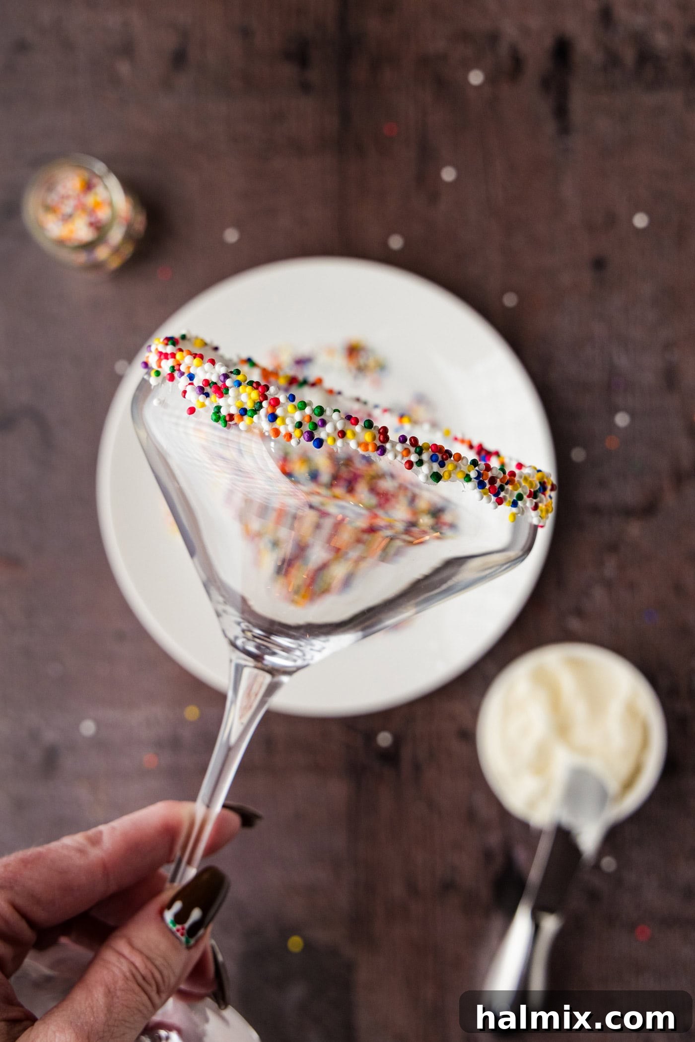 A hand gently coating the rim of a martini glass with vanilla frosting, then rolling it in colorful festive sprinkles for a decorative touch.