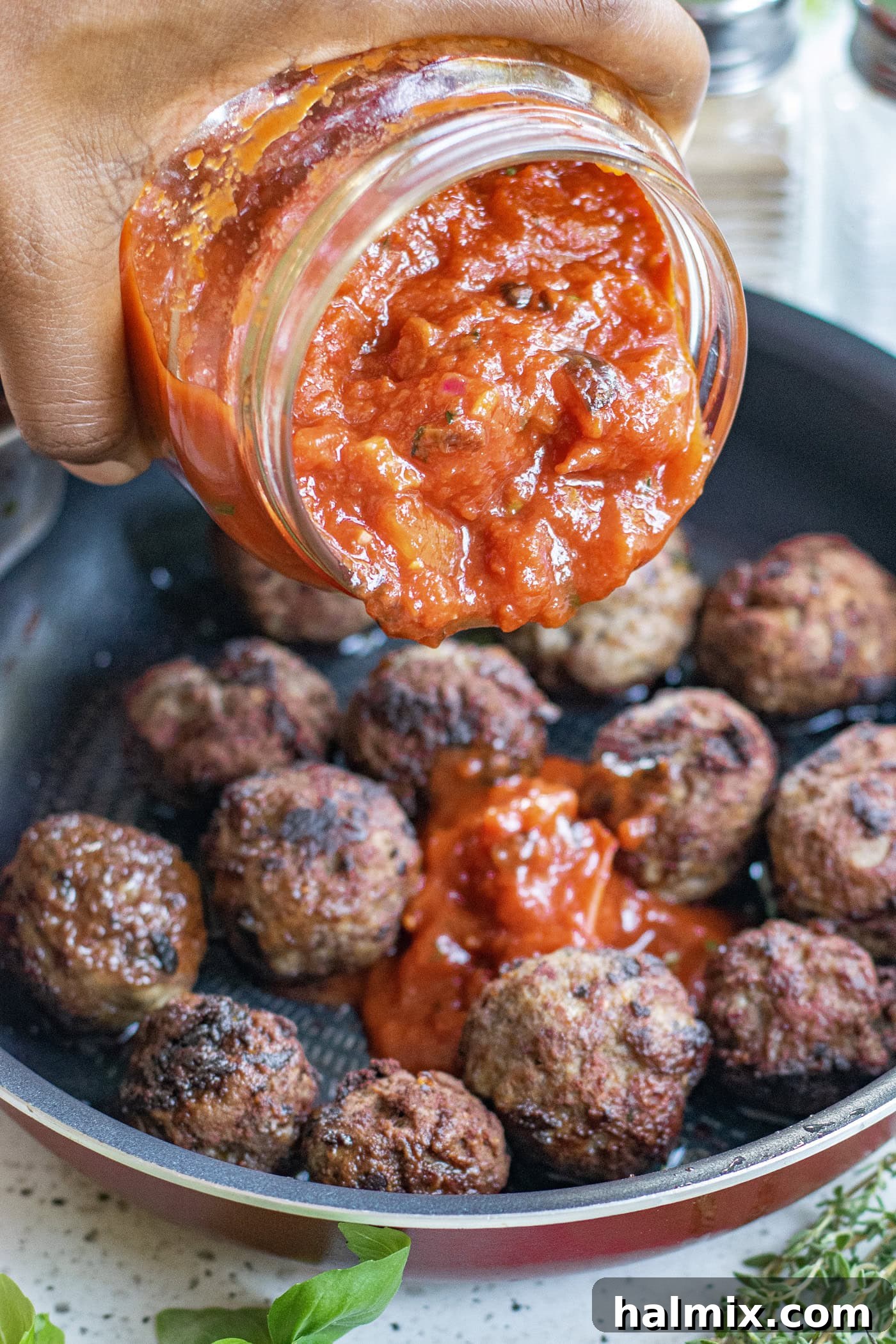 A hand pouring homemade marinara sauce from a jar over a skillet of tender meatballs, ready for a delicious meal.