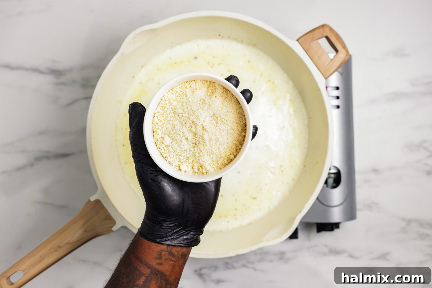 A hand holding a bowl of grated Parmesan cheese over a skillet containing a simmering garlic cream sauce.